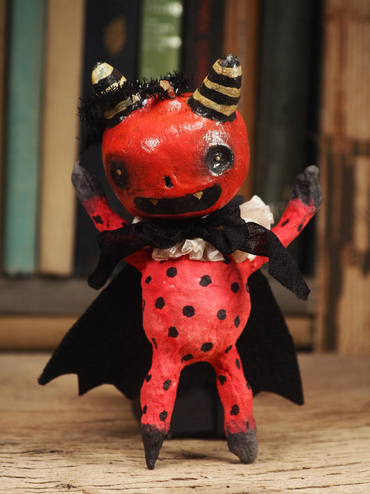 A handmade red and black bat devil doll with black horns, a black cape, and a ruffled white collar, standing on a wooden surface with a blurred background of books. Original hand made Spun cotton Art Doll by Idania Salcido AKA Danita Art.