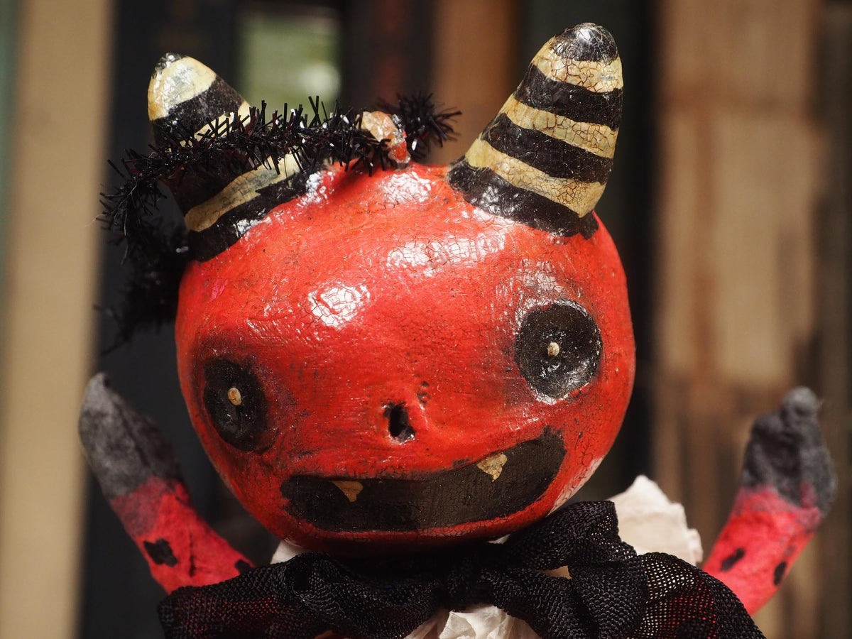 A handmade red and black bat Halloween devil doll with black horns, a black cape, and a ruffled white collar, standing on a wooden surface with a blurred background of books. Original hand made Spun cotton Art Doll by Idania Salcido AKA Danita Art.