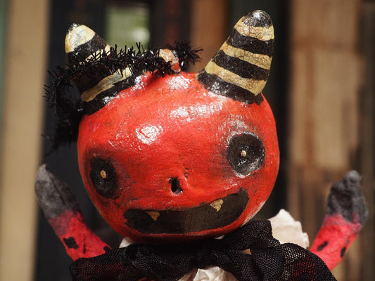 A handmade red and black bat Halloween devil doll with black horns, a black cape, and a ruffled white collar, standing on a wooden surface with a blurred background of books. Original hand made Spun cotton Art Doll by Idania Salcido AKA Danita Art.