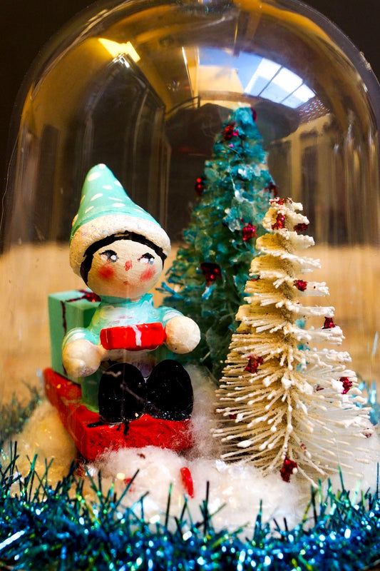 An original handmade christmas tree ornament by Idania Salcido. A clear domed diorama with a holiday scene with Santa Claus elves, Christmas Trees, Elves, Gifts and magical glitter deer.