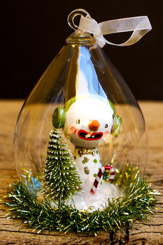 An original handmade christmas tree ornament by Idania Salcido. A clear domed diorama with a holiday scene with Santa Claus elves, Christmas Trees, Elves, Gifts and magical glitter deer.