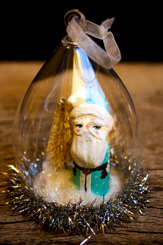 An original handmade christmas tree ornament by Idania Salcido. A clear domed diorama with a holiday scene with Santa Claus elves, Christmas Trees, Elves, Gifts and magical glitter deer.