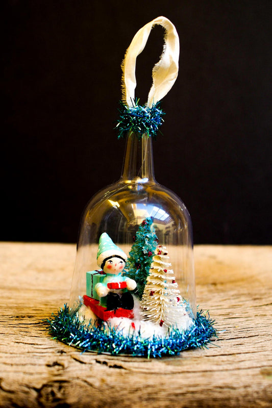 An original handmade christmas tree ornament by Idania Salcido. A clear domed diorama with a holiday scene with Santa Claus elves, Christmas Trees, Elves, Gifts and magical glitter deer.
